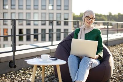 Femme en hijab travaillant sur un ordinateur portable dans un fauteuil bean bag sur une terrasse extérieure.